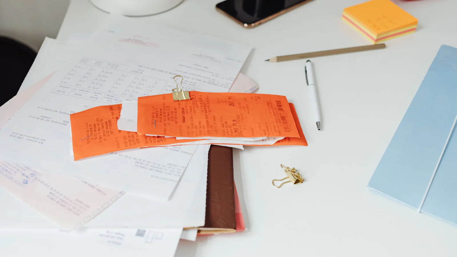 Orange receipts clipped together on top of papers on a white desk, next to a pen and sticky notes