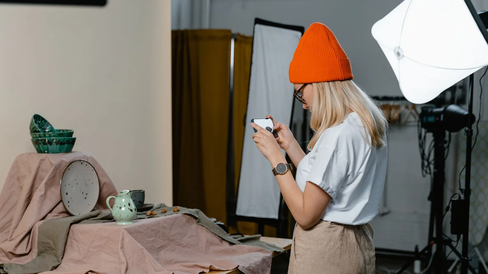 Woman in an orange beanie photographing ceramic bowls and a jug with her phone in a small studio
